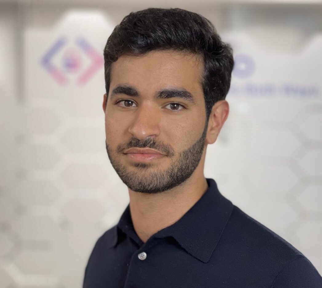 A man with short dark hair and a trimmed beard, wearing a black collared shirt, stands in front of a blurred background featuring geometric patterns and CyberWeek 2024 logos.