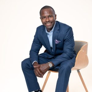 A man in a blue suit sits on a wooden chair, smiling at the camera against a plain white background, ready to share top tips for avoiding digital impersonation and cyber fraud scams.