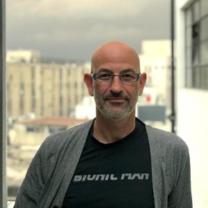 A bald man with glasses and a grey beard stands outdoors in a "Bionic Man" t-shirt and grey cardigan, with buildings and a cloudy sky behind him—perfect for the Tech Success Stories Podcast featuring Memcyco.